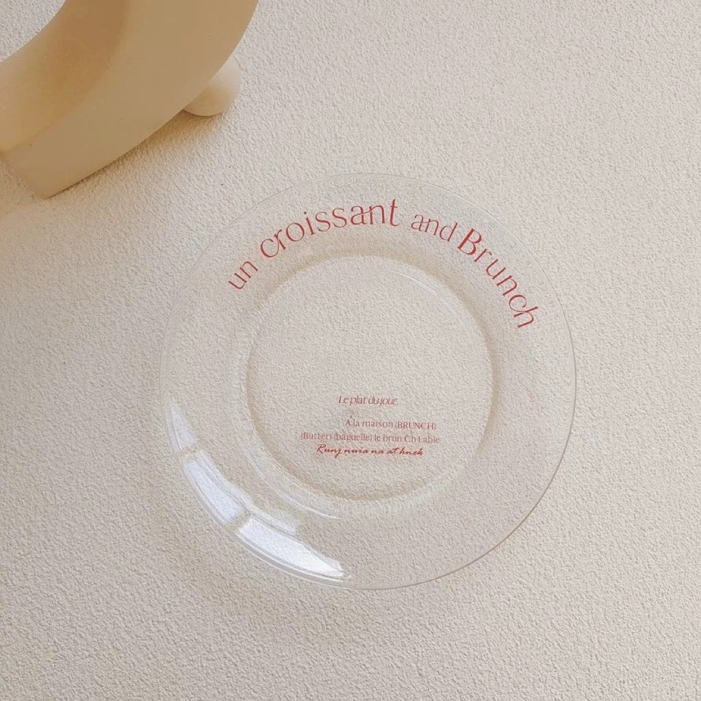 A clear circular glass plate with red printed text reading 'un croissant and Brunch' and smaller text below, photographed on a textured off-white surface with part of a beige object visible in the top-left corner.