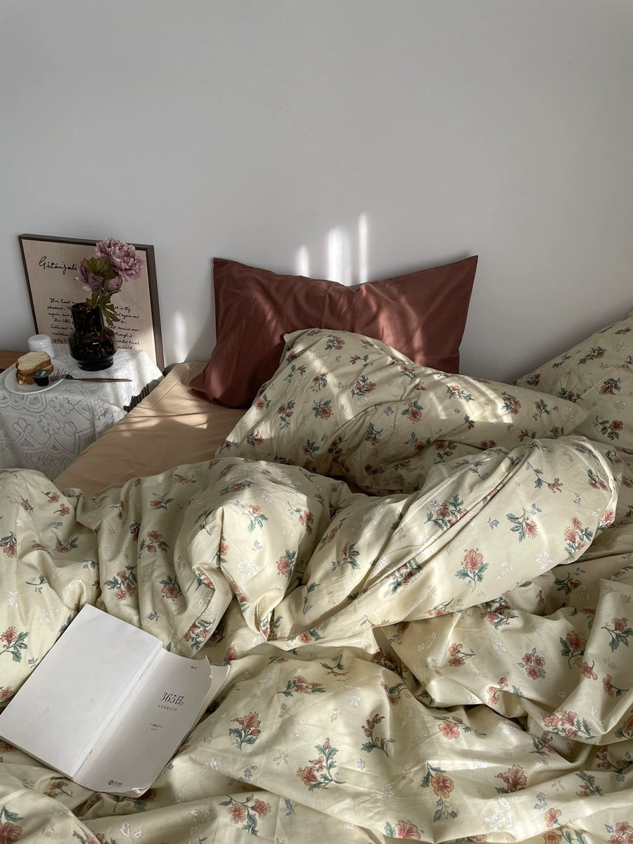 Vintage botanical duvet spread across a sunlit bed with rust pillows, flowers, and an open book.