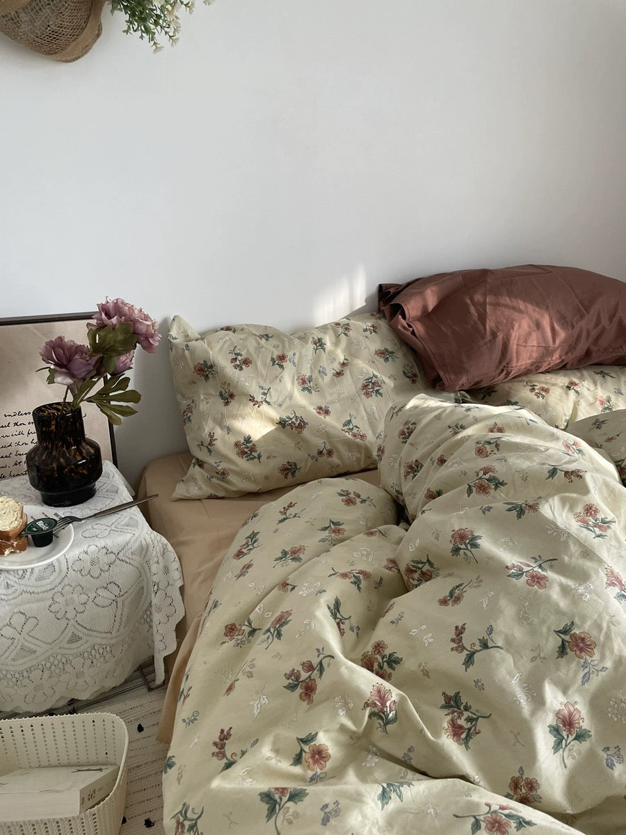 Close view of vintage floral bedding beside rust pillows, flowers, framed art, and a lace-covered table.