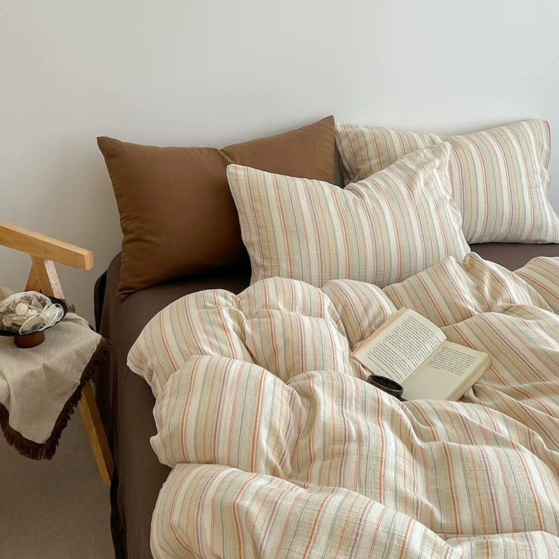 A cozy bed styled in a minimalist bedroom setting, featuring a cream-colored duvet cover and striped pillows with thin red, green, and beige vertical stripes, alongside solid brown accent pillows. An open book and a small wooden chair are visible on the bed and to the side.