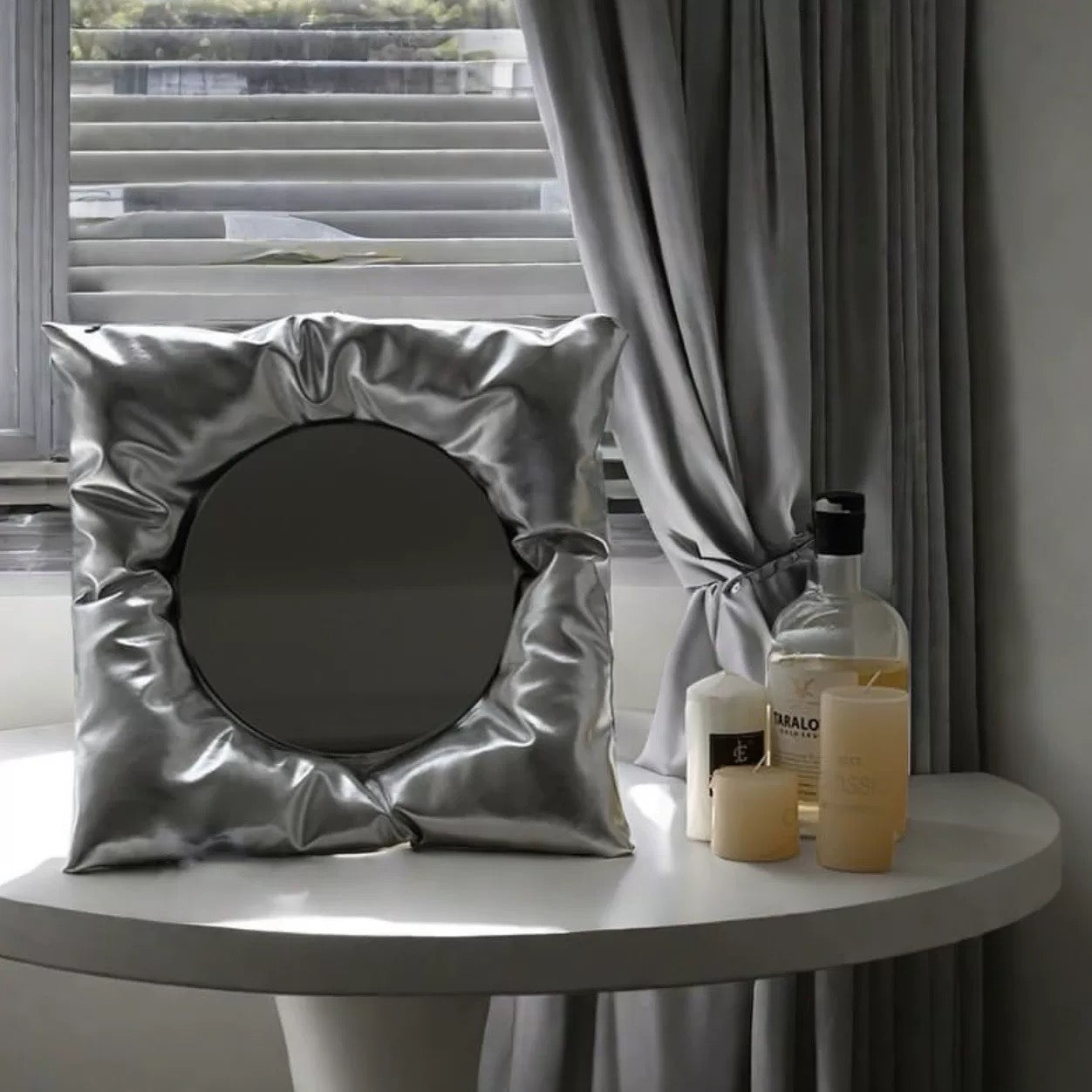 Silver cushion with a round mirror on a table next to candles and bottles against a window backdrop.