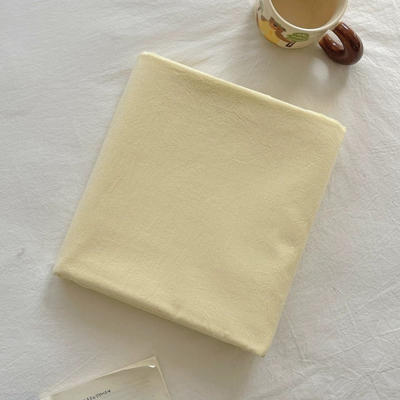 A pale cream-colored fabric pouch or small bag laid flat on a white textured surface, with a ceramic mug partially visible in the top right corner.