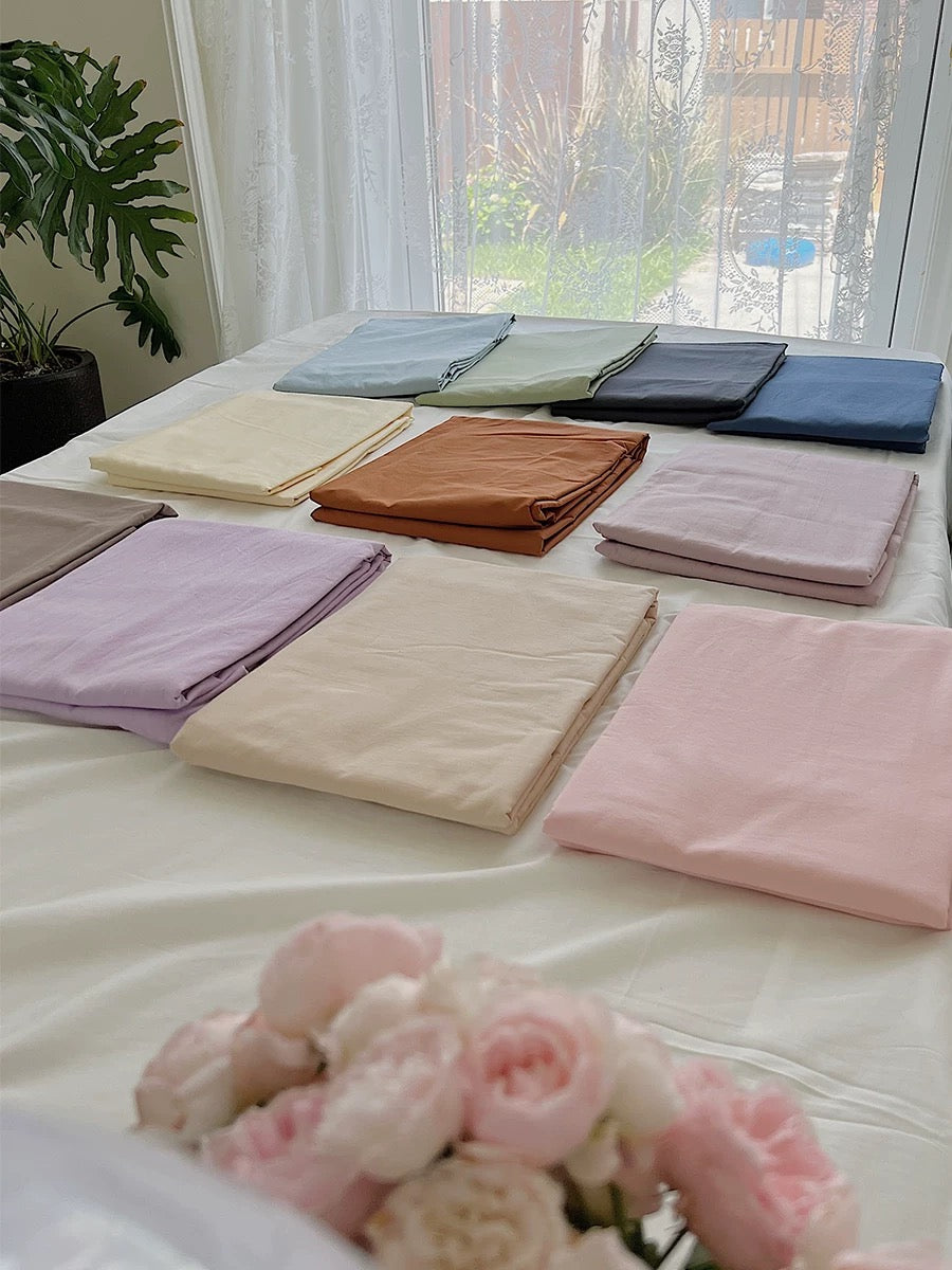 Folded solid color bed sheet options in blue, pink, green and cream are arranged across a white bed.