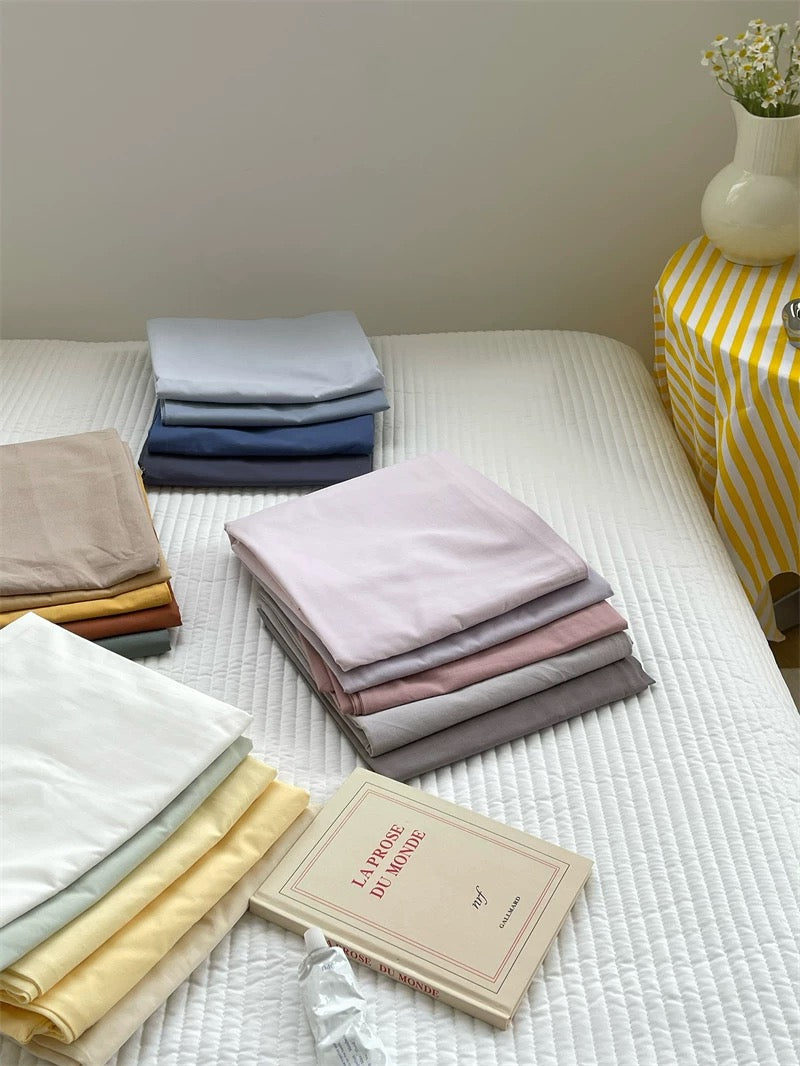 Stacks of folded washed cotton bedding sit on a white bed beside a book and a yellow striped table.