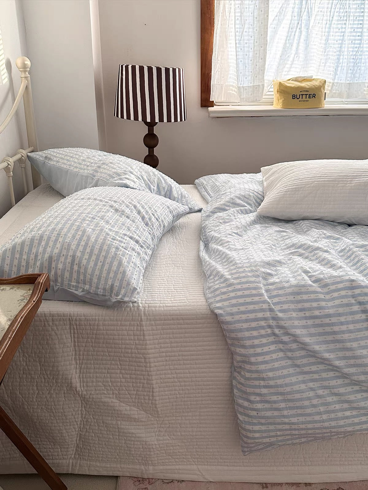 A bed dressed in light blue and white striped bedding, including pillows, a duvet cover, and a textured cream sheet, styled in a cozy bedroom setting with a striped lamp and a window.