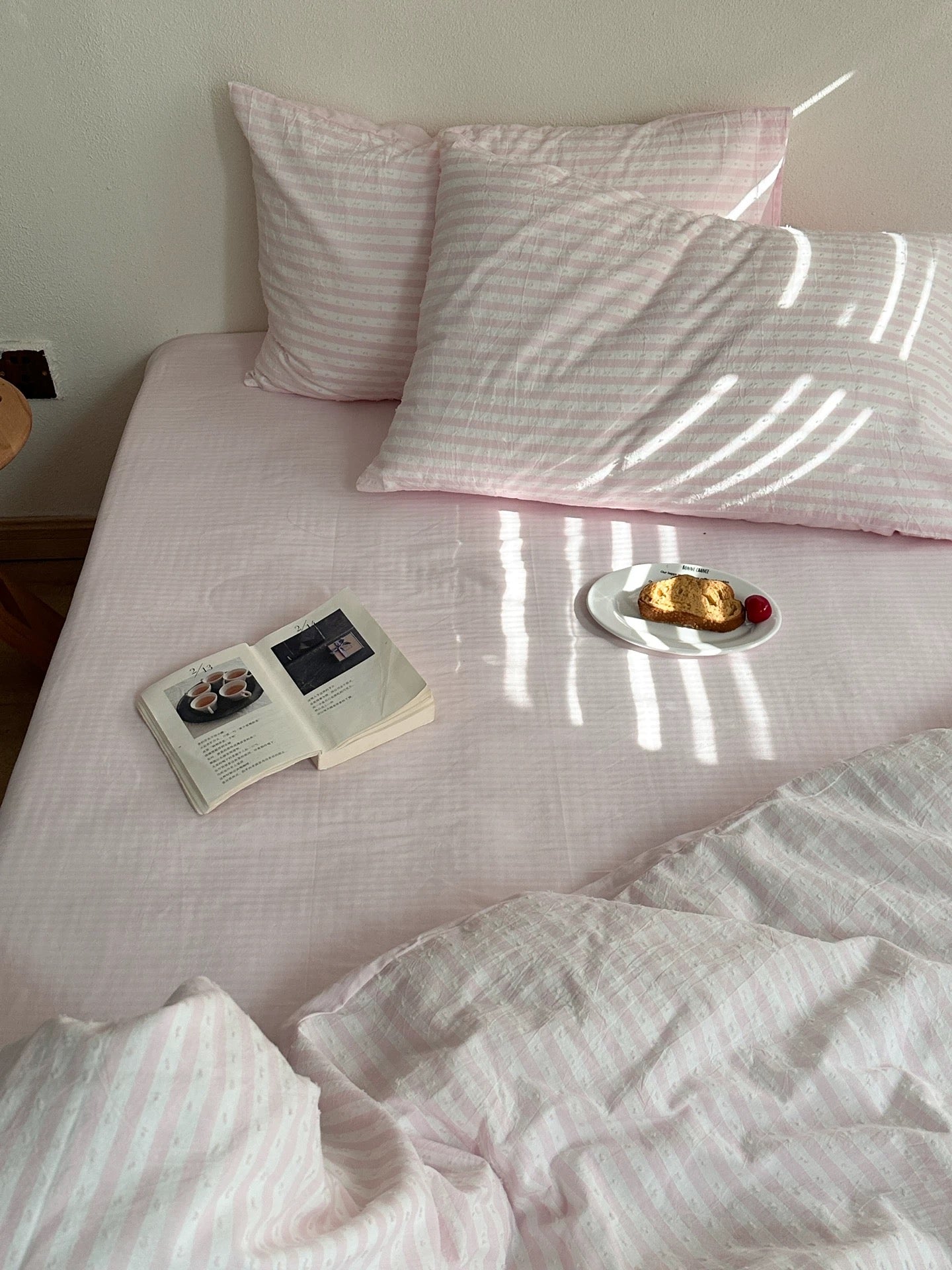 Pink and white striped bedding set on a bed, featuring two pillows, a matching sheet, and a duvet, styled with an open book and a small plate of food in a softly lit room.