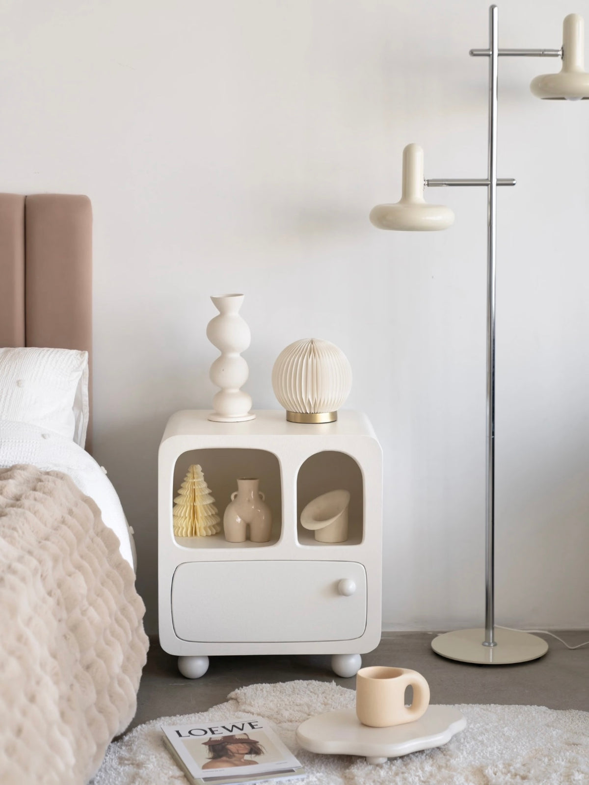 A white bedside cabinet stands beside a bed, with two open compartments above its closed lower drawer.