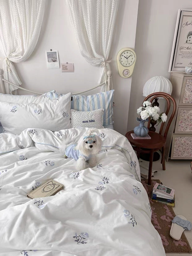 Dog in pajamas on a bed with floral bedding, next to a nightstand with a clock and lamp.