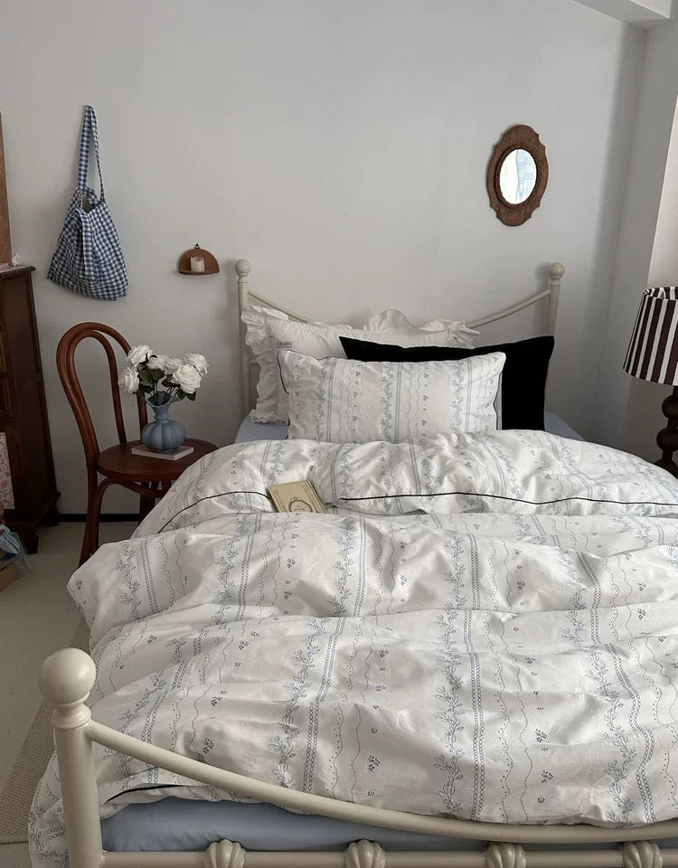 Bedroom with a bed featuring a white and gray patterned comforter, a chair with a bag, and a mirror on the wall.