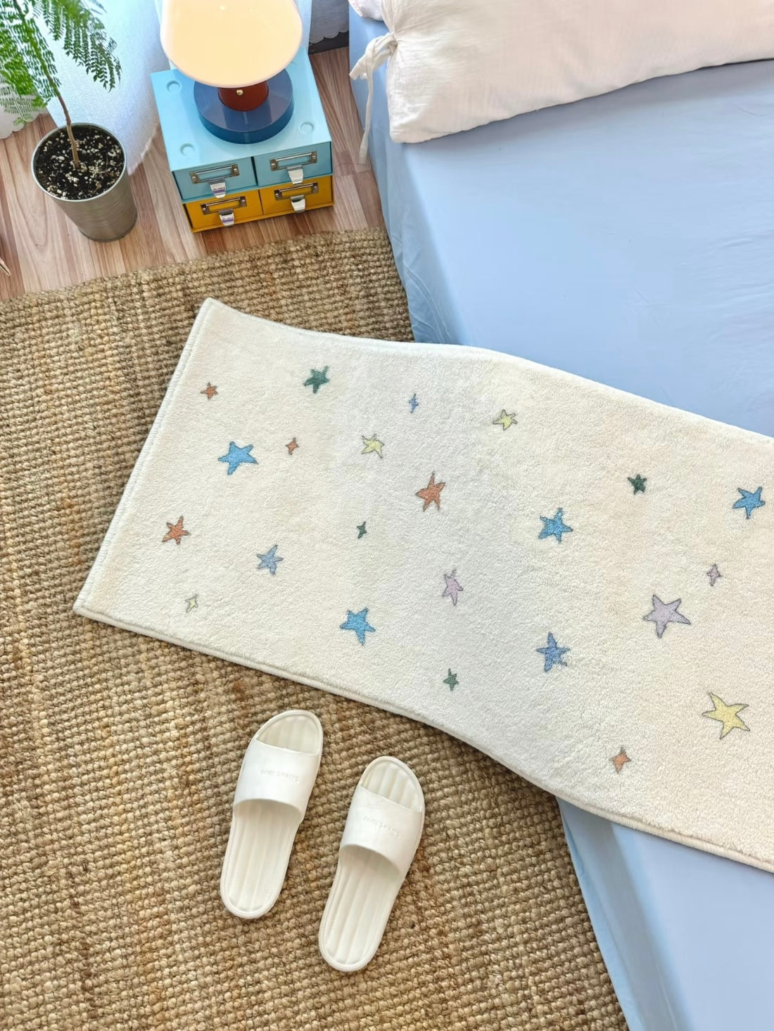 A star rug with blue motifs on a cream background lies beside a blue bed, with slippers at the near edge.