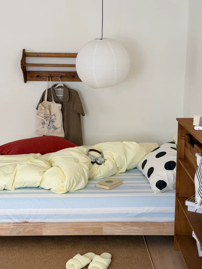 A cozy bedroom scene with a bed made up in pale yellow bedding, a blue-and-white striped sheet, a red pillow, a black-and-white polka dot pillow, a wooden wall rack with a tote bag and brown shirt, a white pendant light, and a wooden nightstand.